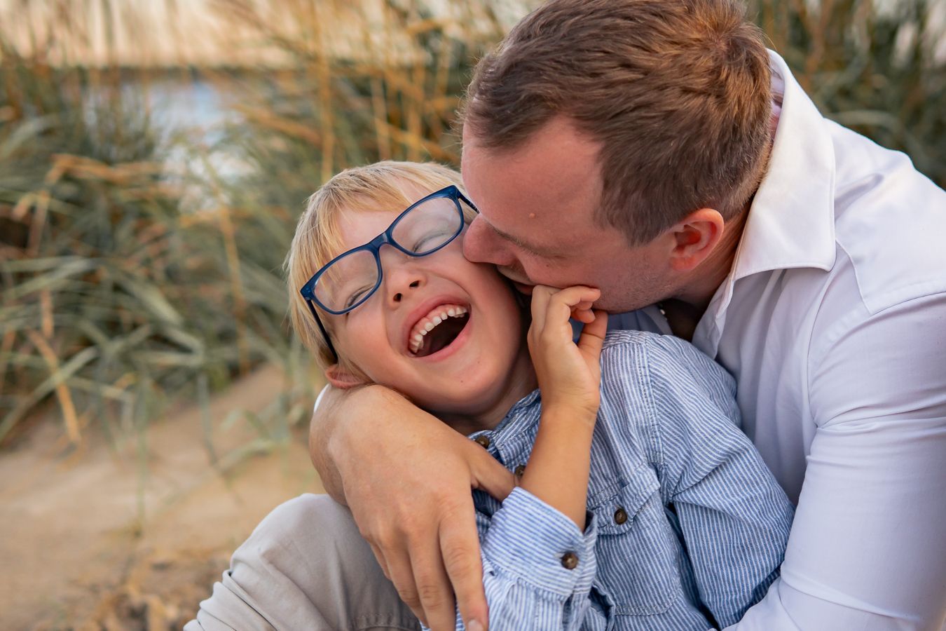 Lekfull stund där pappa pussar sin son som skrattar på sandstranden i solnedgång med högt gräs bakom. Fotograf, familjefotograf, Umeå