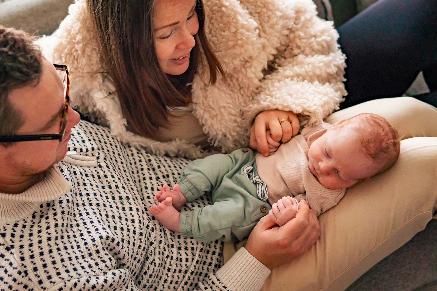 Nyblivna föräldrar tittar på sin sovande nyfödda bebis som ligger i pappas knä och de håller i händerna. Fotograf, nyföddfotograf, Umeå