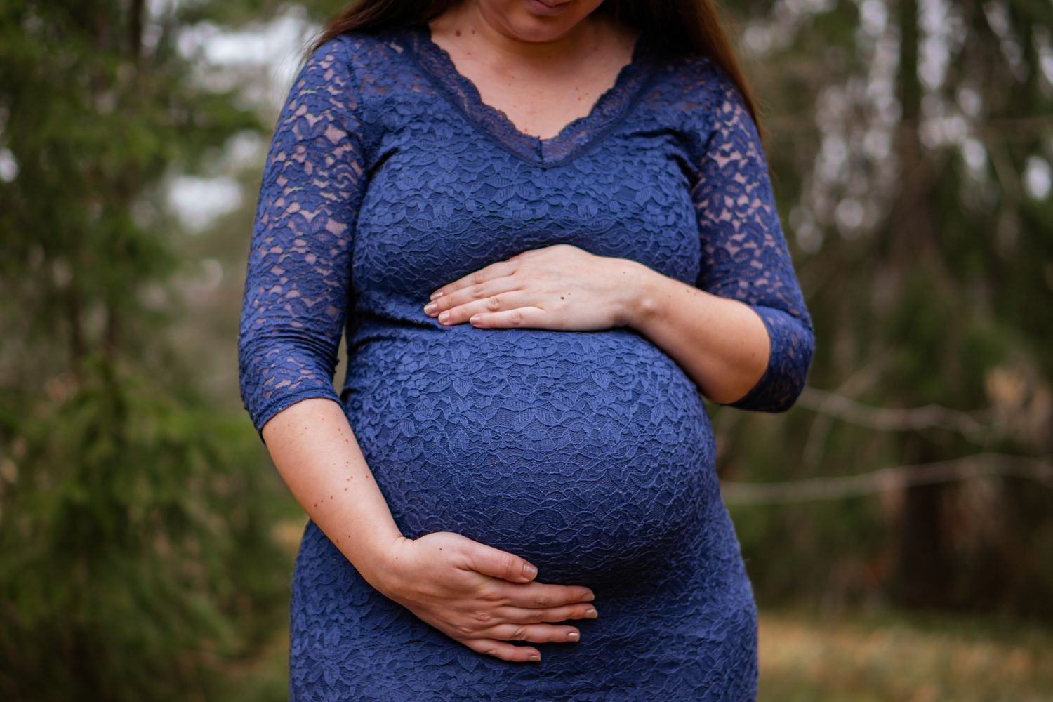 Gravid kvinna i blå spetsklänning håller om sin gravida mage med skogen i bakgrunden. Gravidfotograf, Umeå, Västerbotten