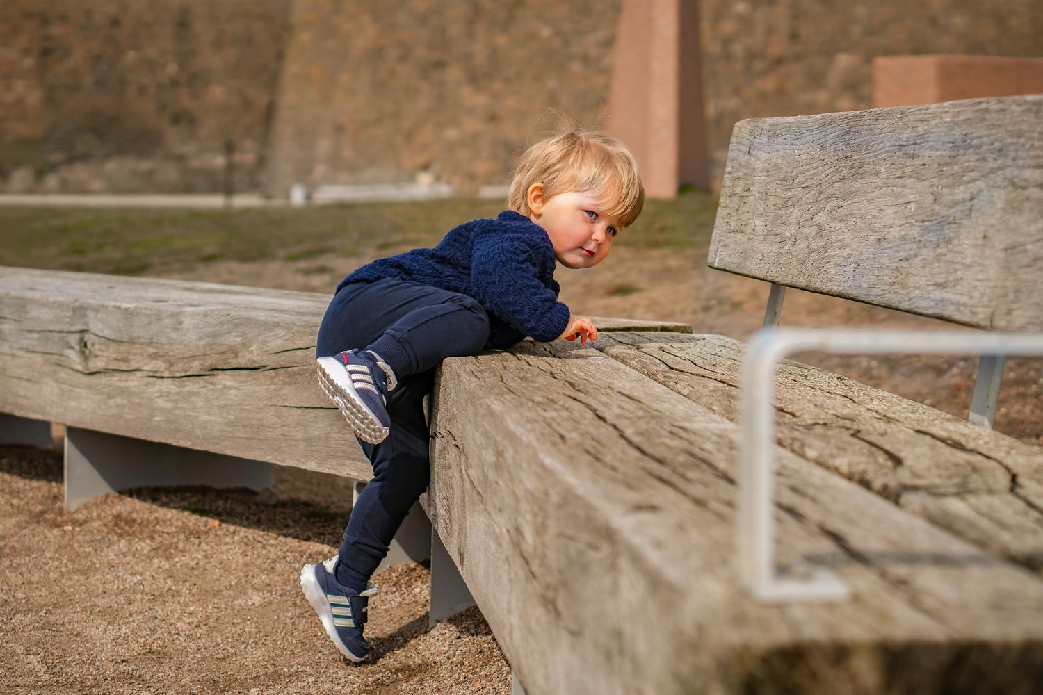Litet barn i marinblå kläder försöker klättra upp på en träbänk utomhus i solen. Familjefotograf, Umeå, Väaterbotten