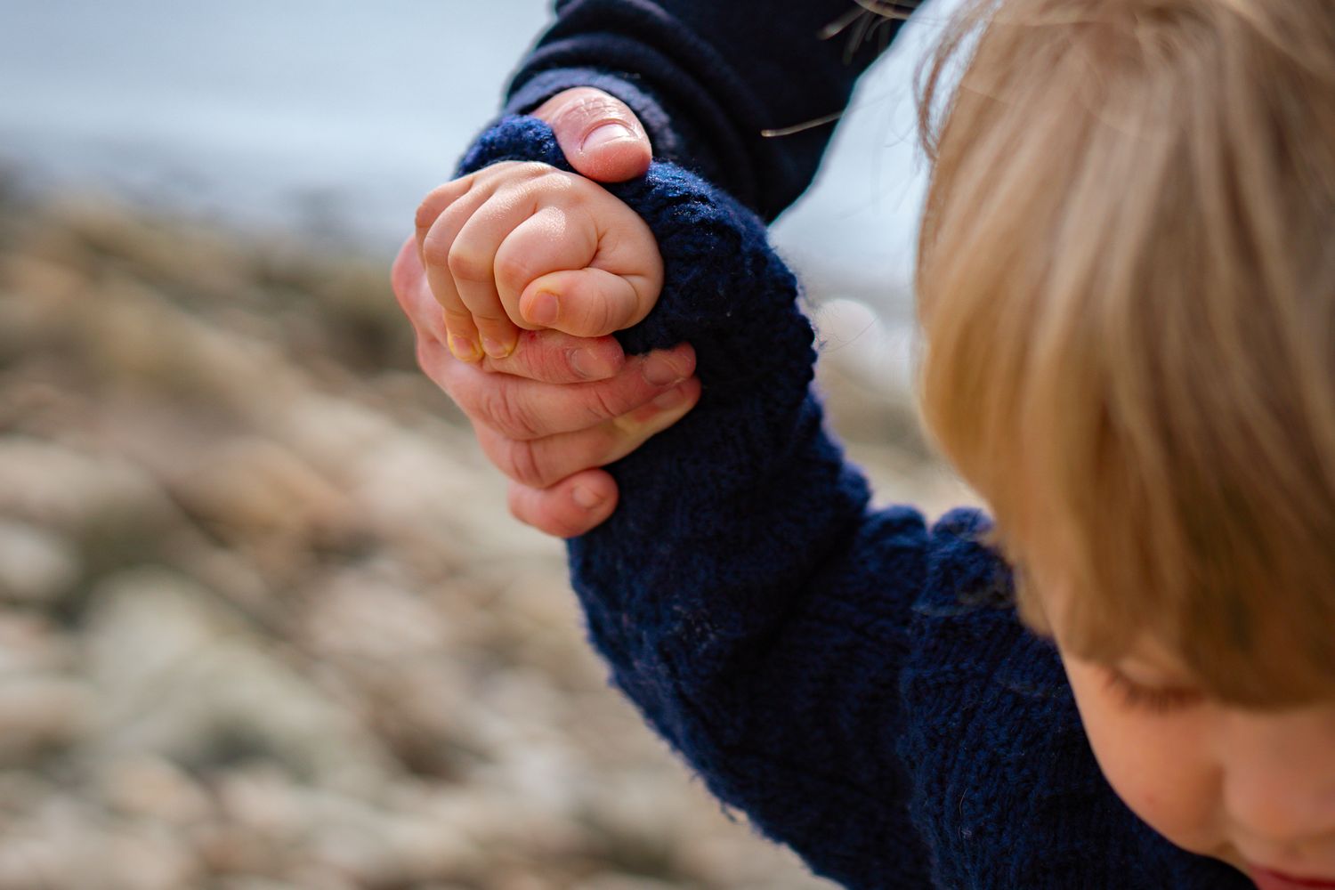 Närbild på en barnhand som stöttas av en vuxen hand för att hålla balansen på en stenstrand. Familjefotograf, Umeå, Västerbotten