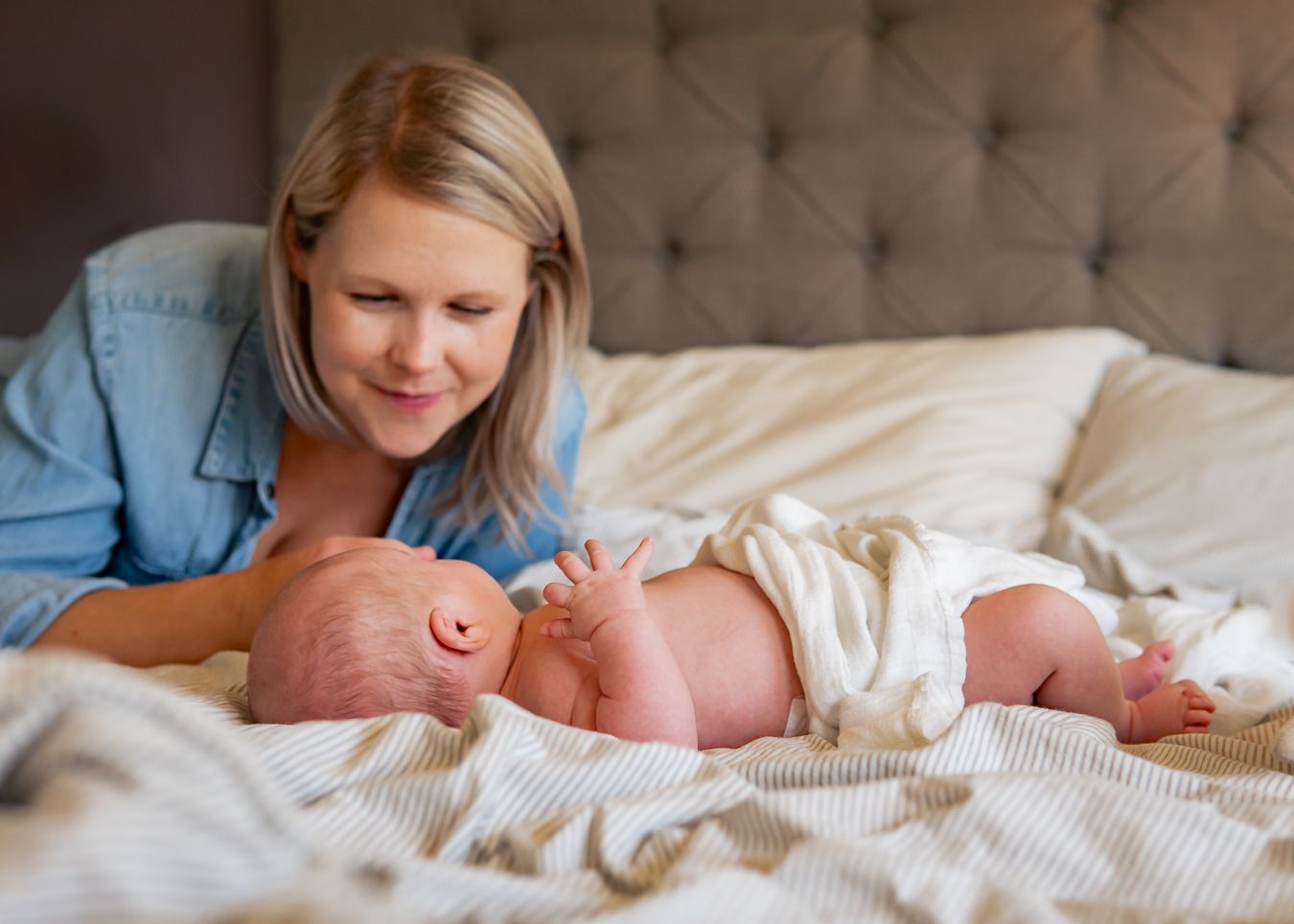 Glad mamma i denim skjorta ler kärleksfullt mot sin bebis i sängen. Fotograf, Nyföddfotograf, Umeå, Västerbotten