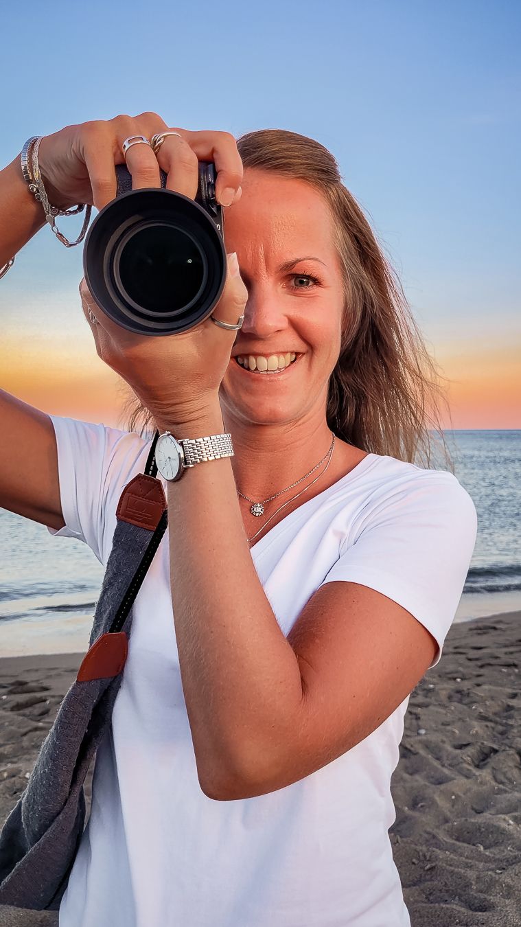 Fotograf håller upp systemkameran, kikar fram bakom den och ler stort på en strand i solnedgången. Fotograf, Umeå, Västerbotten