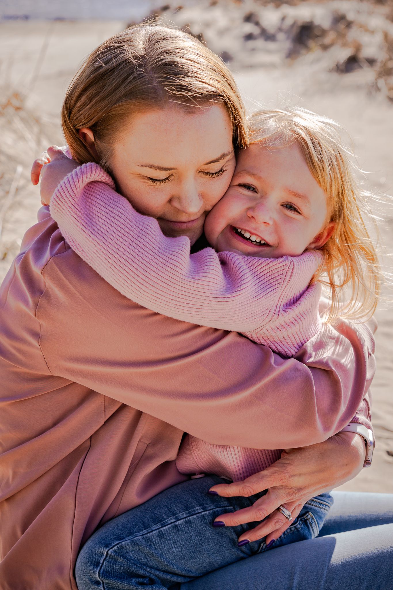 Kärleksfull bild på mamma som kramas ordentligt av dotter i famnen med armarna runt halsen på sandstranden en solig dag. Fotograf, Umeå