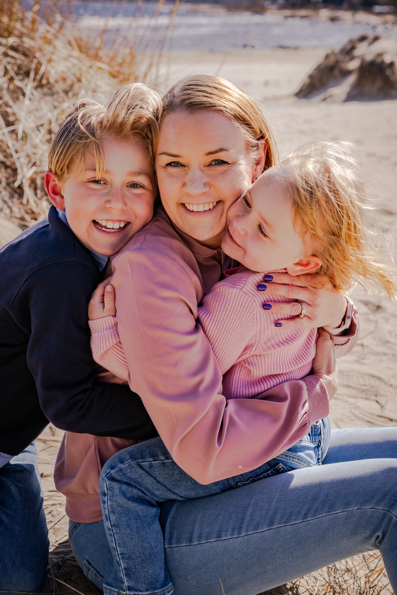 En kärleksfull närbild på mamma och hennes barn som sitter tätt och skrattar på en sandstrand en solig dag. Fotograf, familjefotograf, Umeå