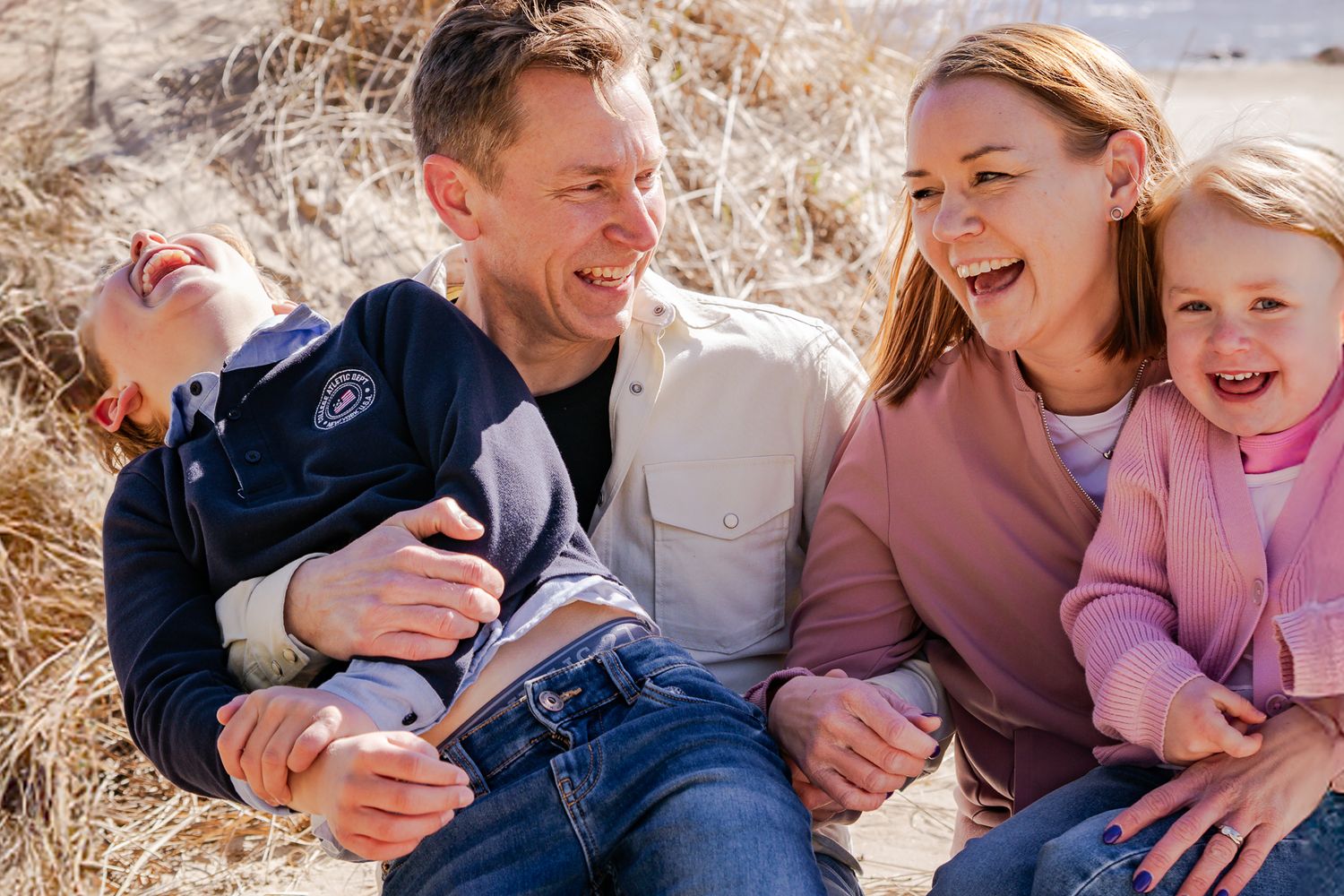 En familj med två barn sitter tillsammans och skrattar stort vid sanddynorna på stranden en solig dag. Fotograf, familjefotograf, Umeå
