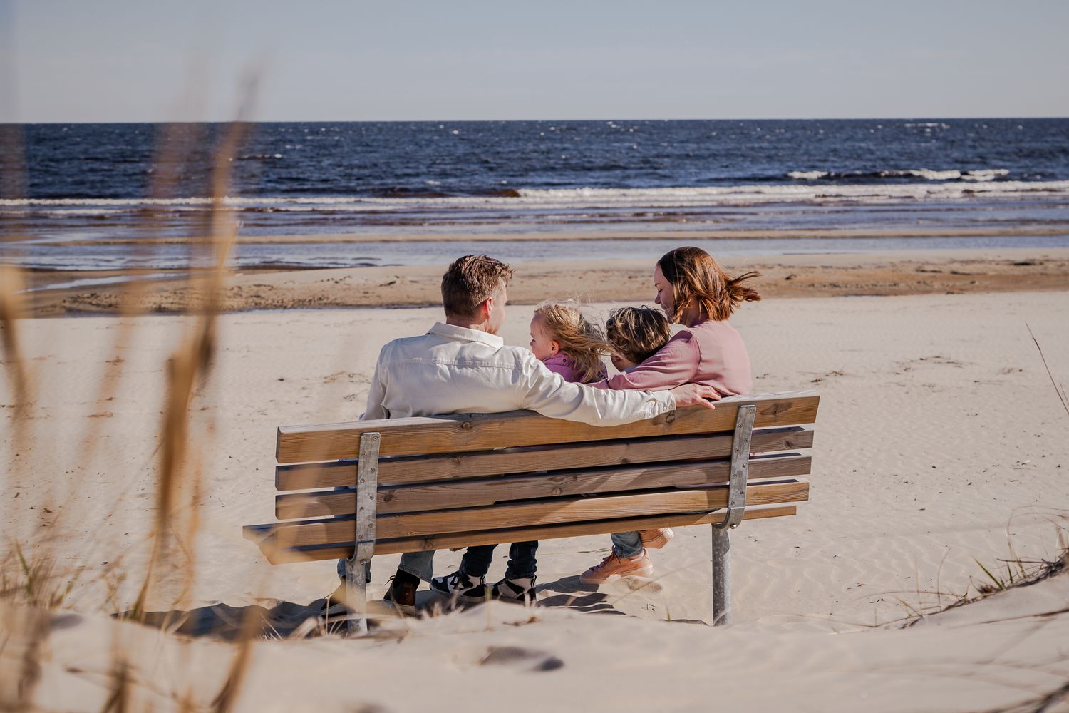 Familj sitter tillsammans på en träbänk på sandstranden med vinden i håret och havet framför sig. Fotograf, familjefotograf, Umeå