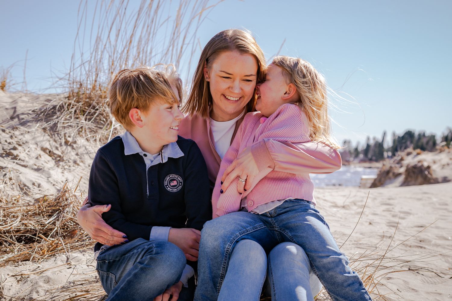 En mamma sitter med sina två barn nära sig i en stund av kärlek och glädje på sandstranden en solig dag. Fotograf, familjefotograf, Umeå