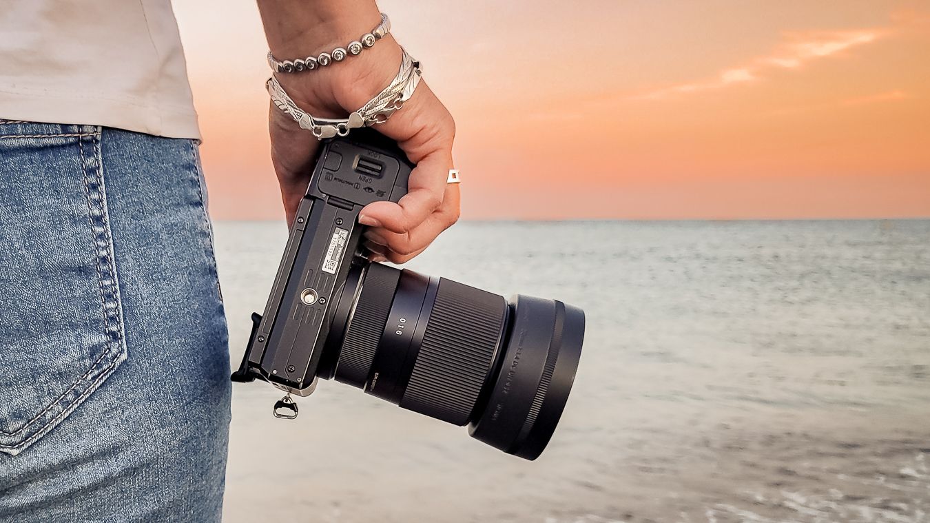 Närbild på en hand med silverarmband som håller en systemkamera med havet och solnedgång i bakgrunden. Fotograf, Umeå, Västerbotten