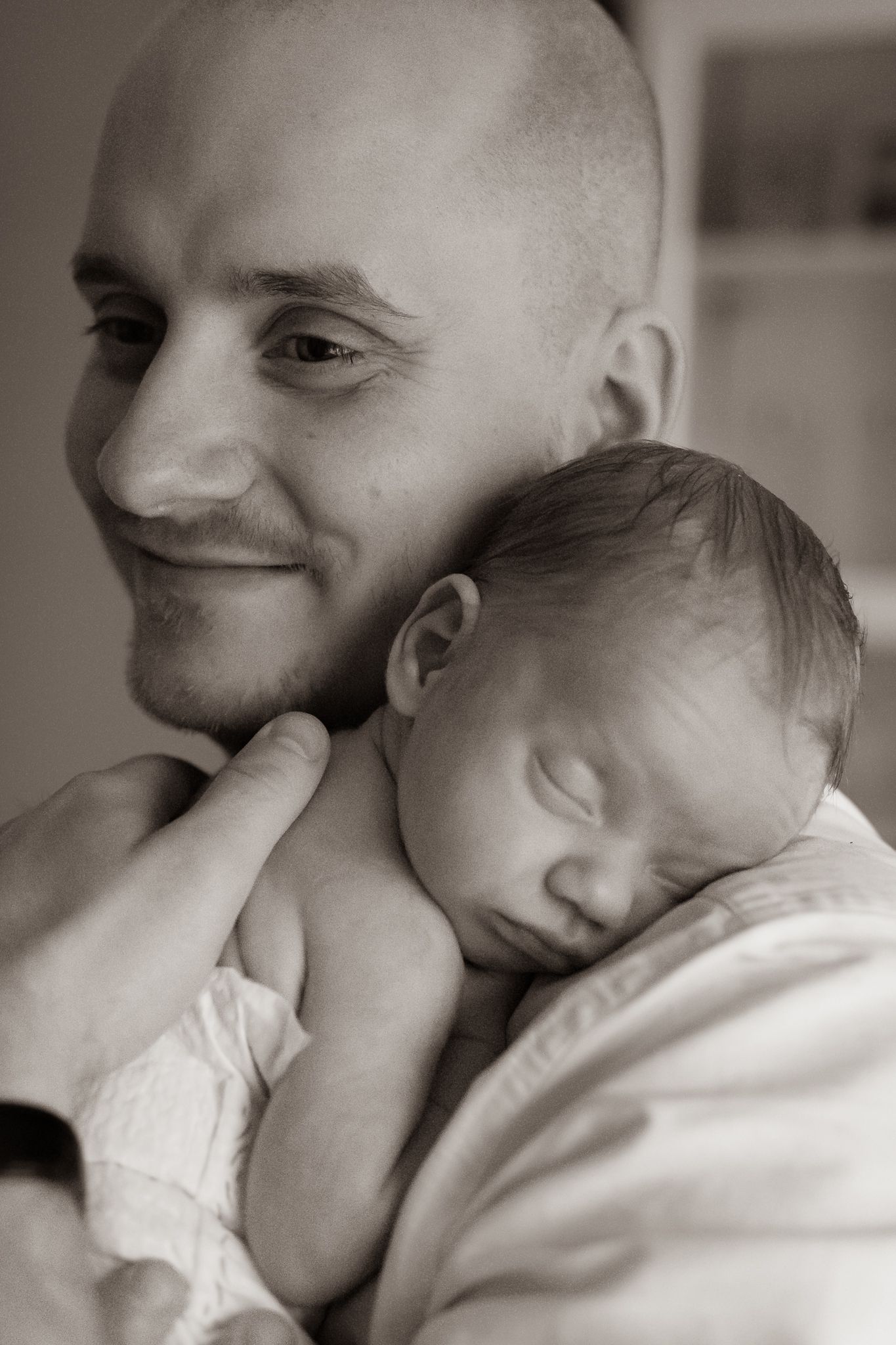 Svartvit bild på nyfödd sovande bebis som vilar mot sin leende pappas axel. Fotograf, nyföddfotograf, Umeå, Västerbotten
