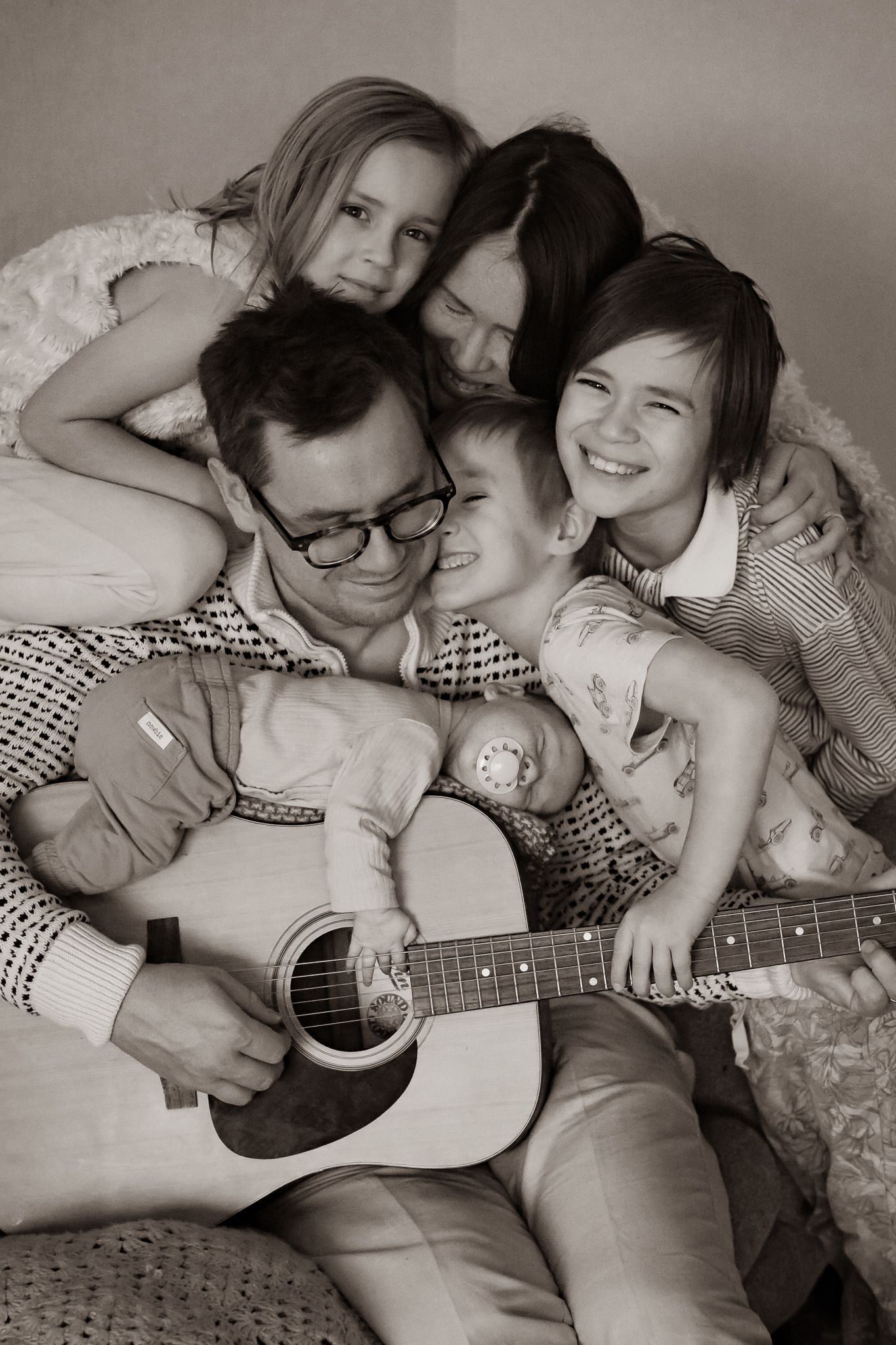 Svartvit bild med en mysig gruppkram med en hel familj i soffan där pappa spelar gitarr och bebis ligger på gitarren. Fotograf, Umeå