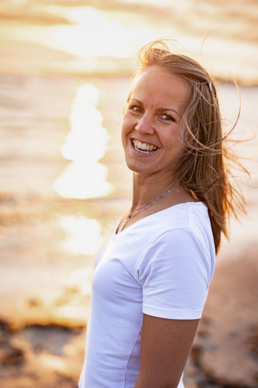 Tjej i vit t-shirt ler stort på en strand i gyllene solnedgång och vinden i håret. Fotograf, Umeå, Västerbotten