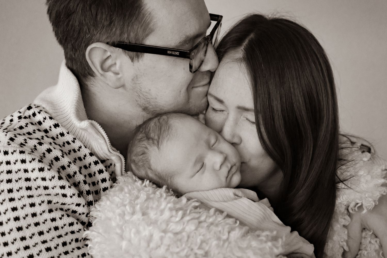Svartvit bild på närheten hos föräldrar med en sovande bebis i famnen, alla tätt intill varandra med ansiktena. Nyföddfotograf, Umeå