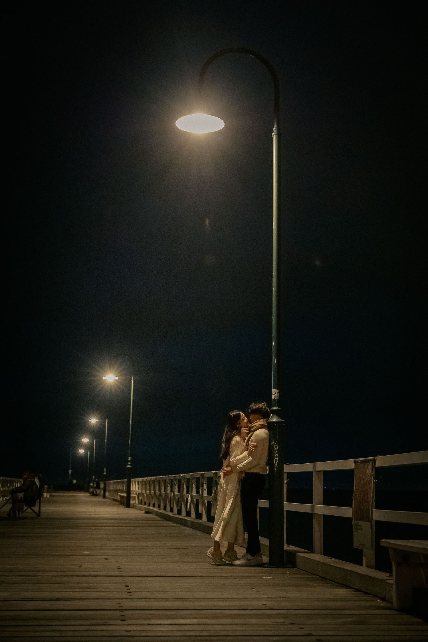 Silhouettes stand on a dimly lit wooden pier at night with glowing street lamps creating a romantic atmosphere.