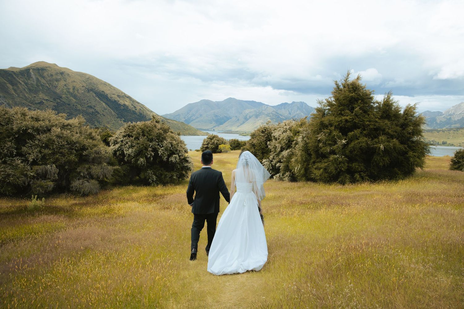 A couple in wedding attire walks through a scenic mountain meadow with golden grass and distant peaks in view.