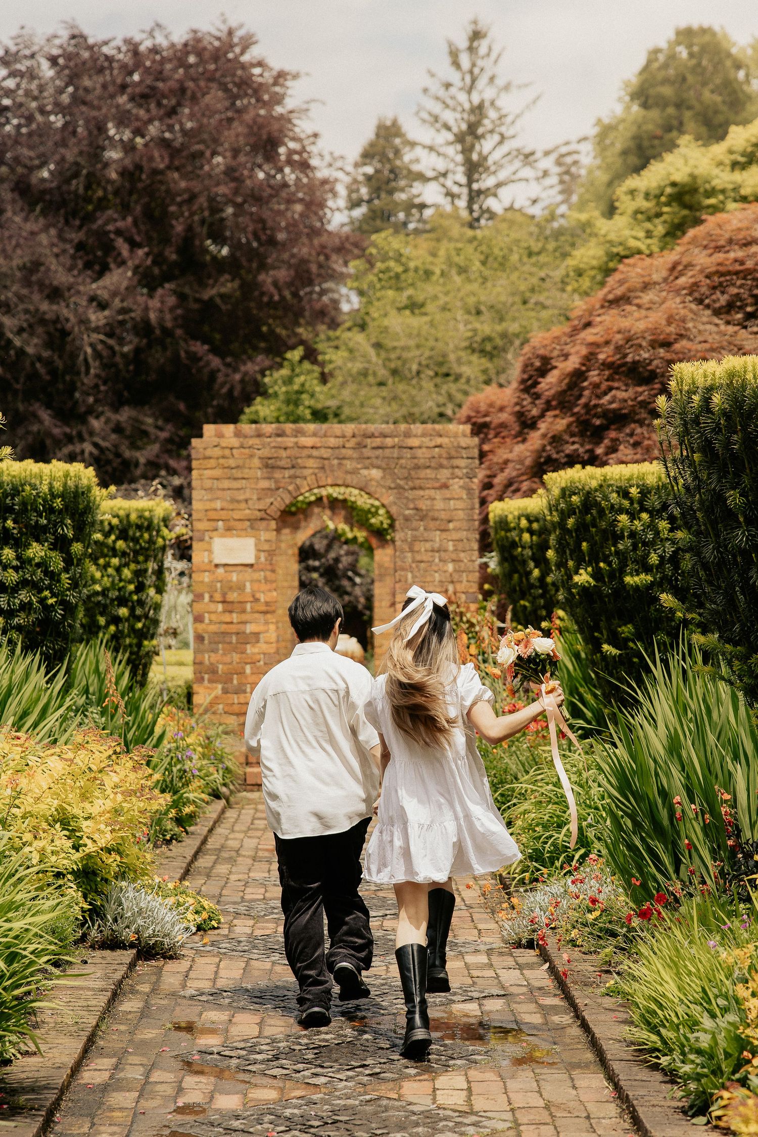 Three figures walk together down a brick path surrounded by lush garden landscaping and flowering plants.