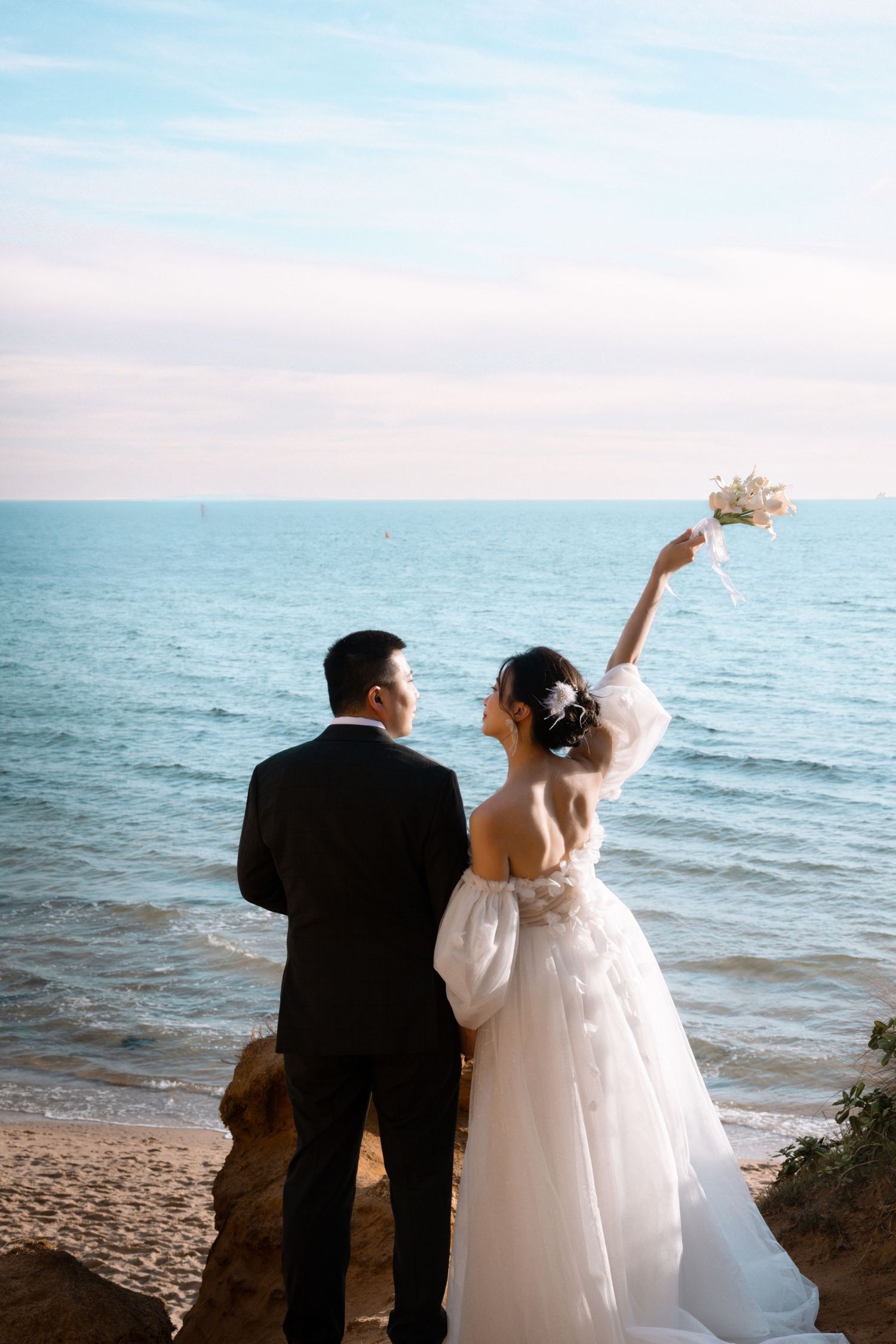 A couple in wedding attire stands at the beach during sunset, throwing something into the ocean.