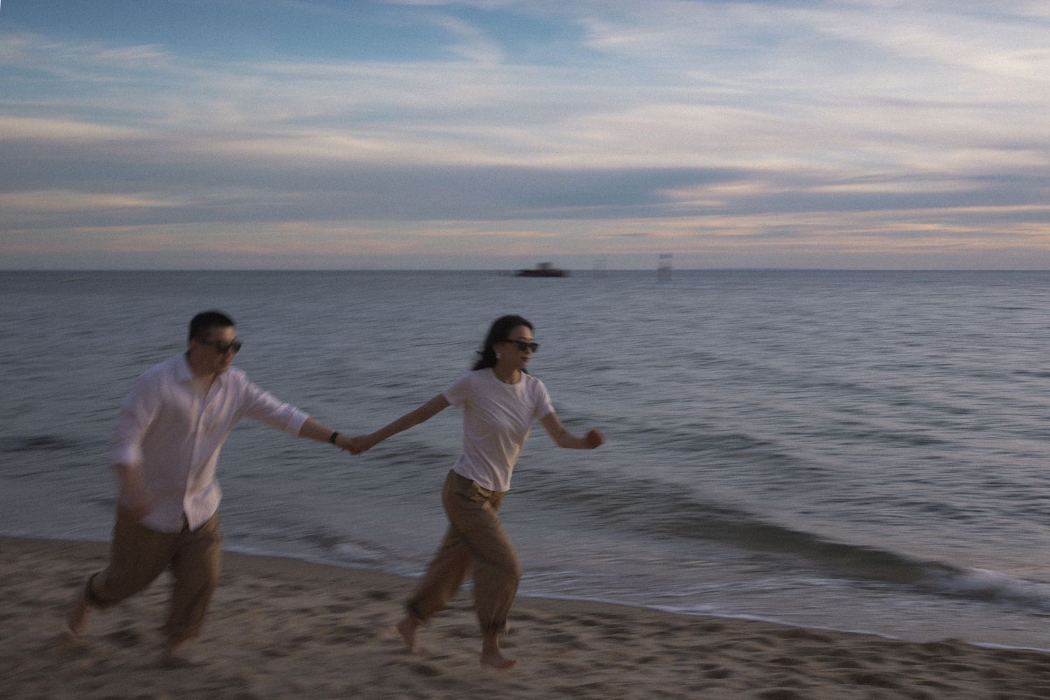 Two people run joyfully together along the beach at dusk with a peaceful ocean and cloudy sky in the background.
