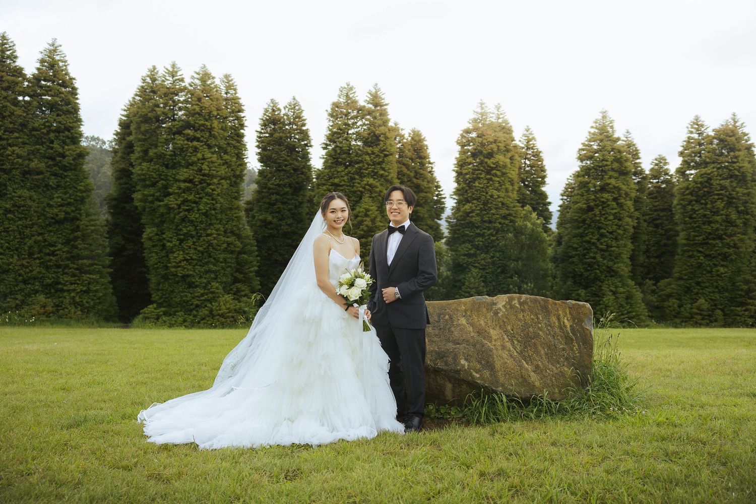A couple in formal wedding attire poses for photos in a scenic evergreen forest setting.