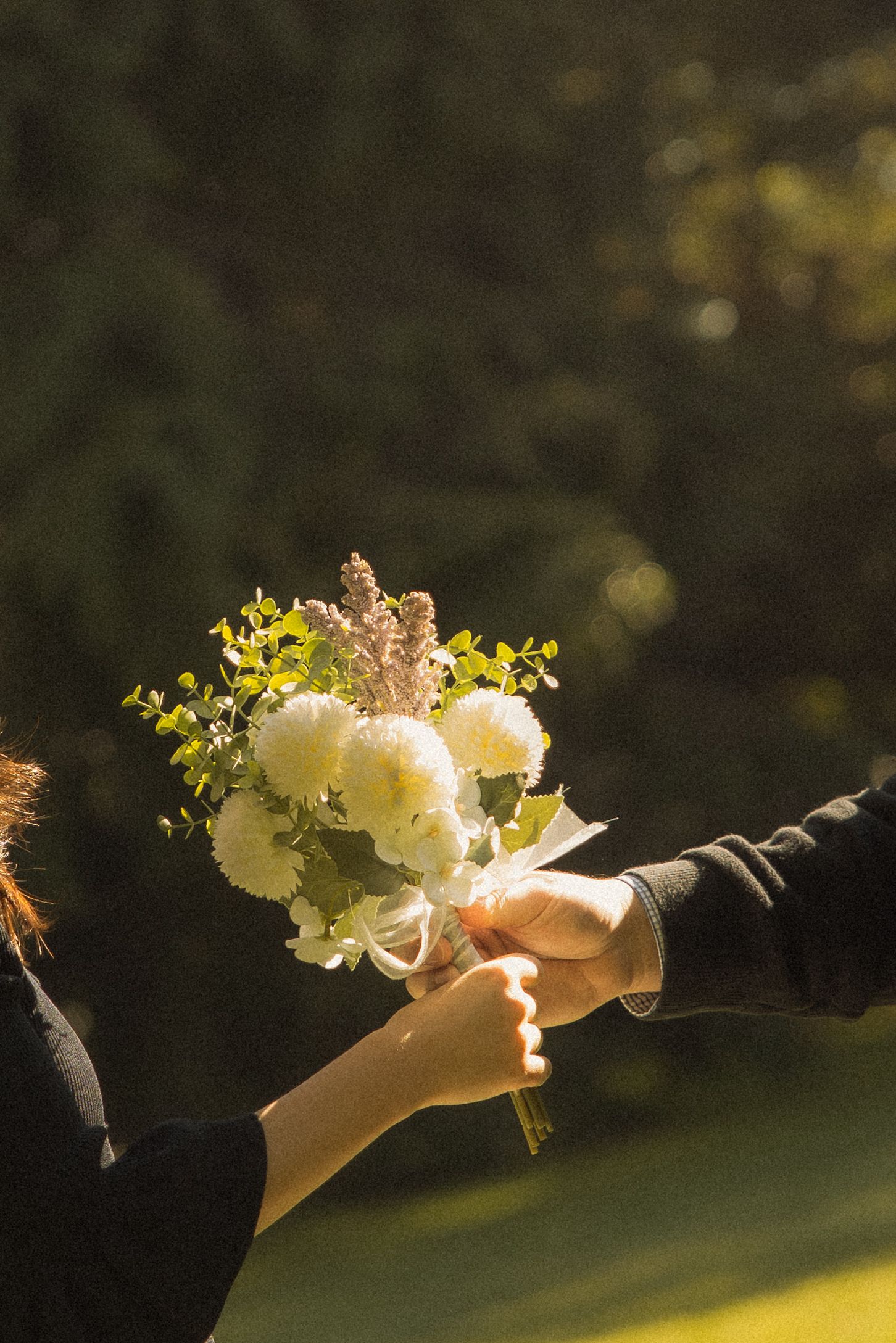 Hands exchanging a beautiful white and yellow floral bouquet in warm sunlight during an outdoor ceremony.