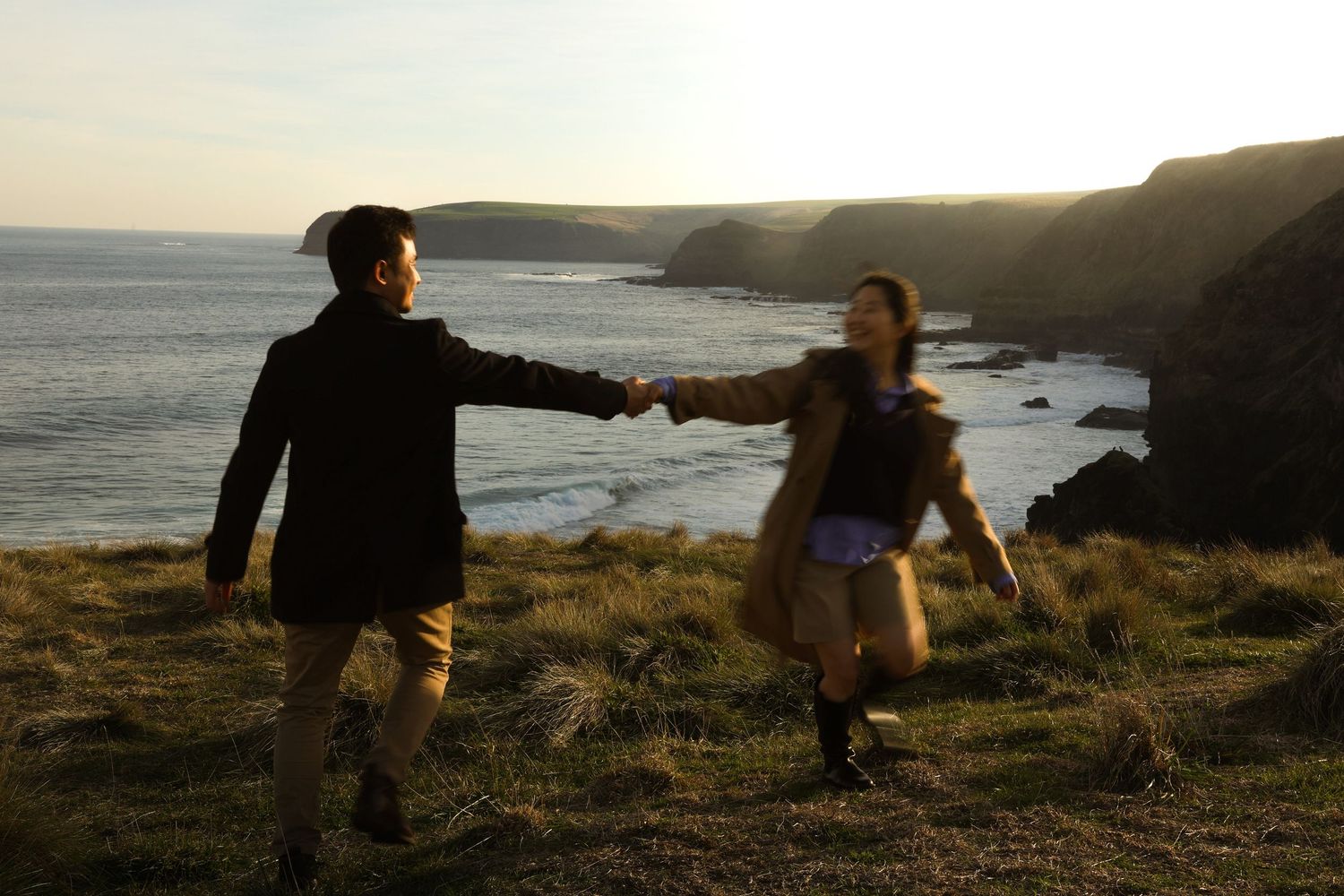 Two people hold hands while walking along a scenic coastal cliff at sunset with ocean views in the background.