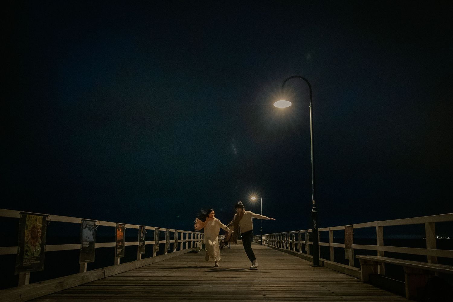 A dark wooden pier extends into the water under a night sky with illuminated lamp posts casting a warm glow.