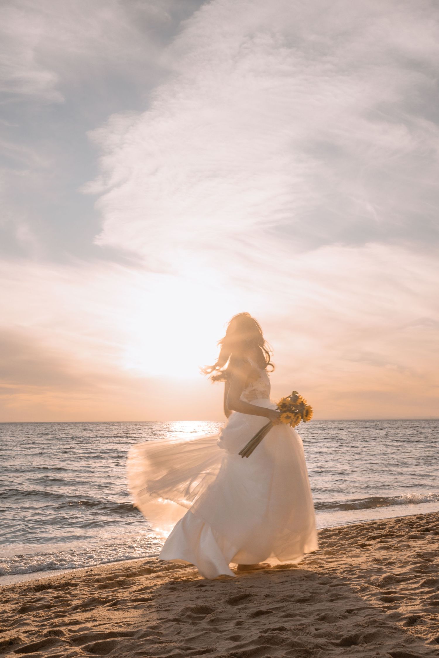 Silhouette of bride in flowing white dress sitting on beach at golden sunset over ocean waves.