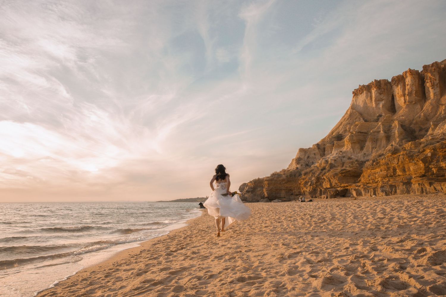 A person walks along a sandy beach at sunset with golden cliffs and ocean waves in the background.