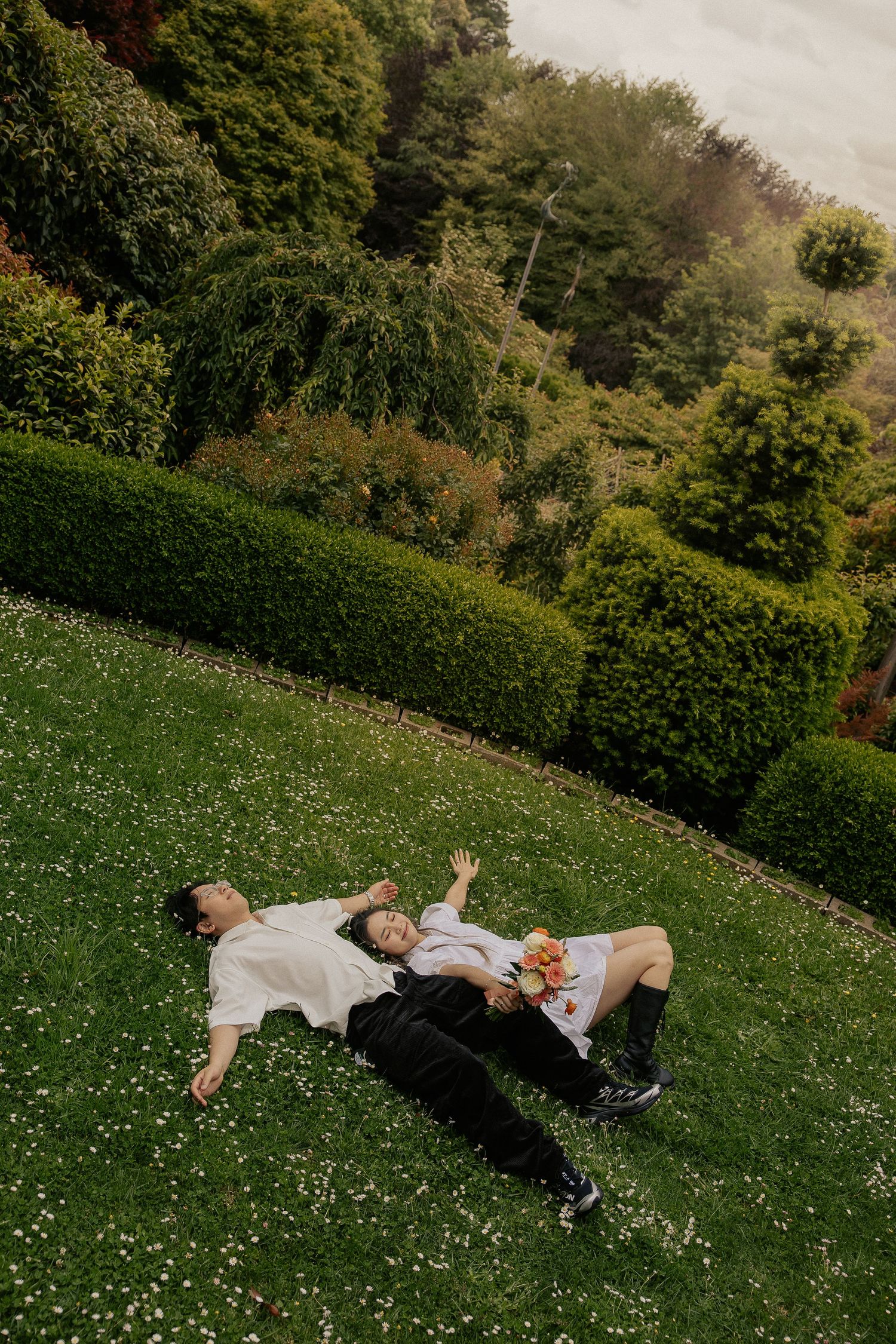 Two people lying on a manicured lawn surrounded by neatly trimmed hedges and lush garden landscaping.