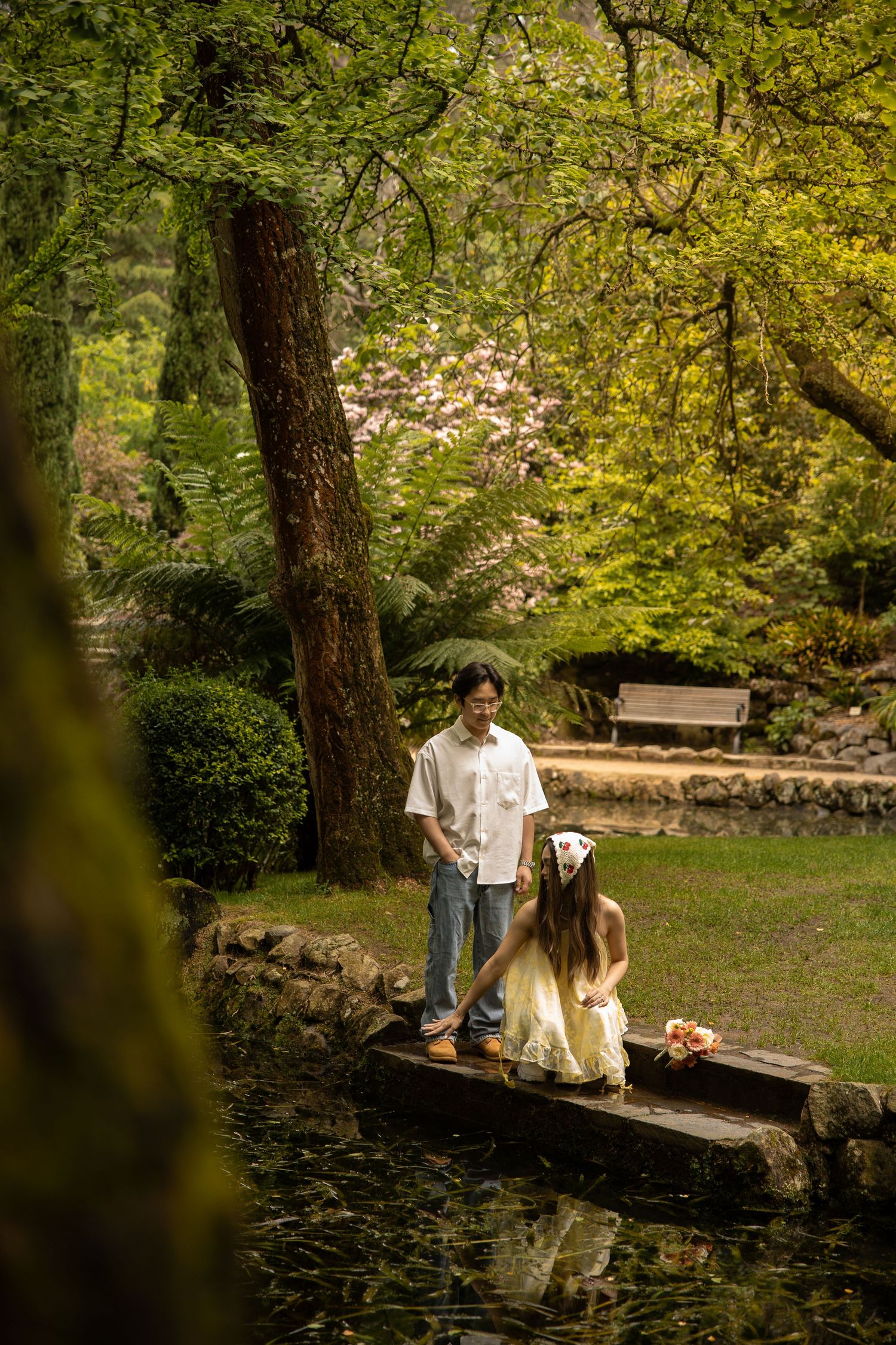 A couple strolls together near a tranquil pond in a lush garden setting with blooming trees and a wooden bench.