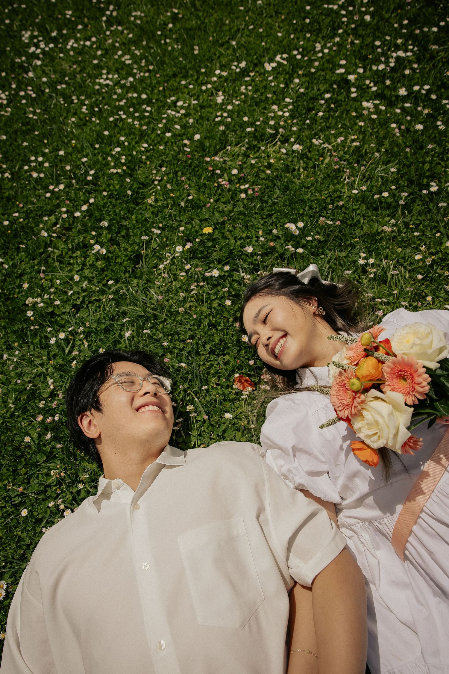 A person in a white dress lies on grass covered in daisies while holding colorful flowers.