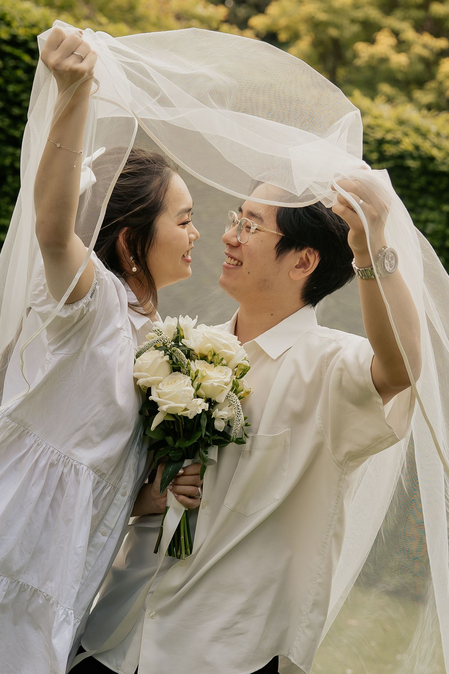 A couple in white attire playfully poses under a flowing veil in an outdoor garden setting.