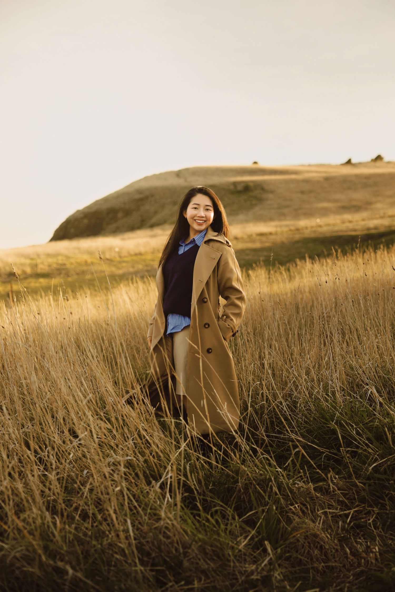 Person in tan coat standing in golden grass field during sunset with rolling hills in background.