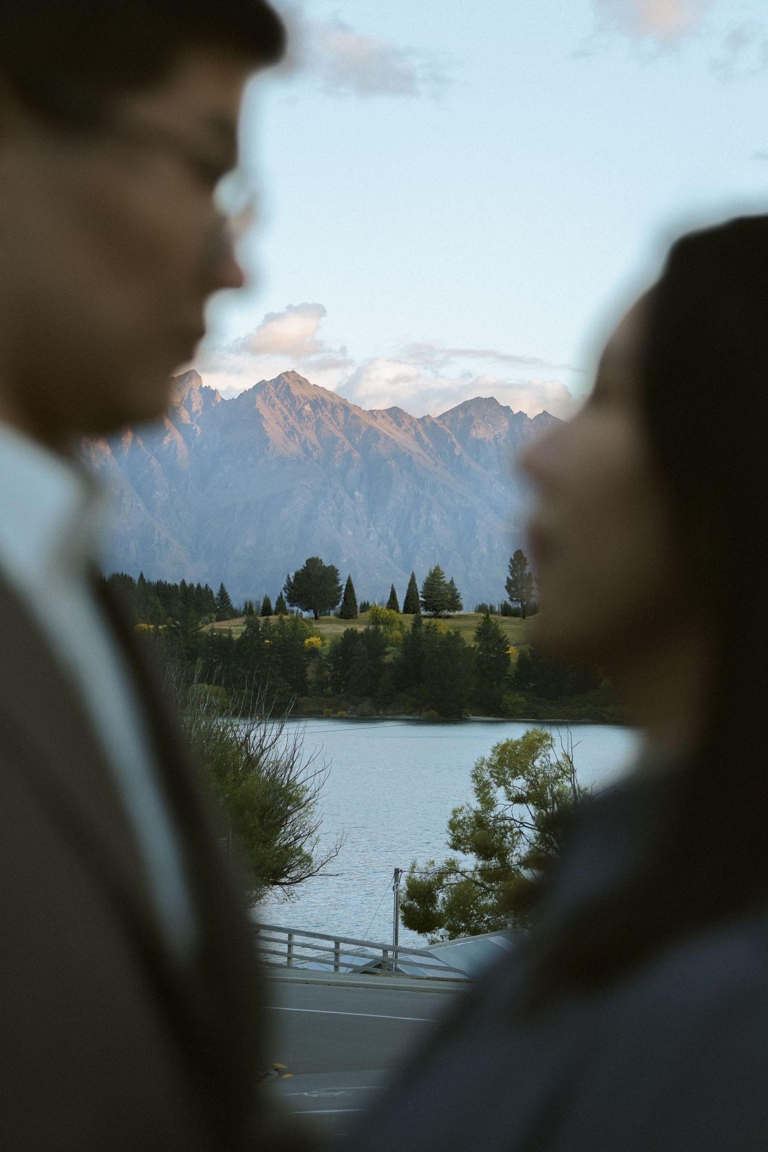 Mountain peaks glow at sunset behind a tranquil lake with trees and a wooden fence in the foreground.