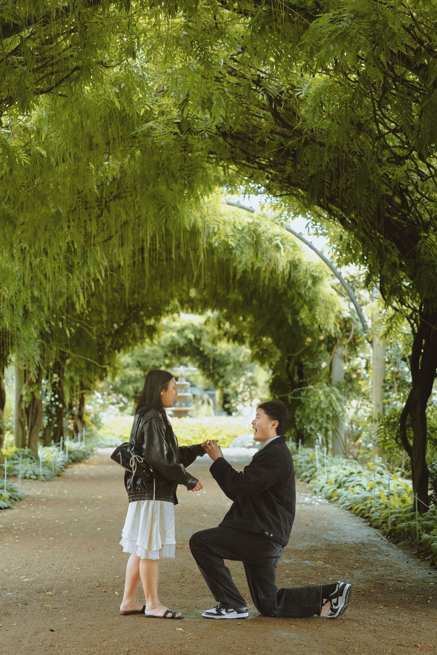 Two people share a quiet moment together under a lush green archway of trees in a tranquil garden setting.