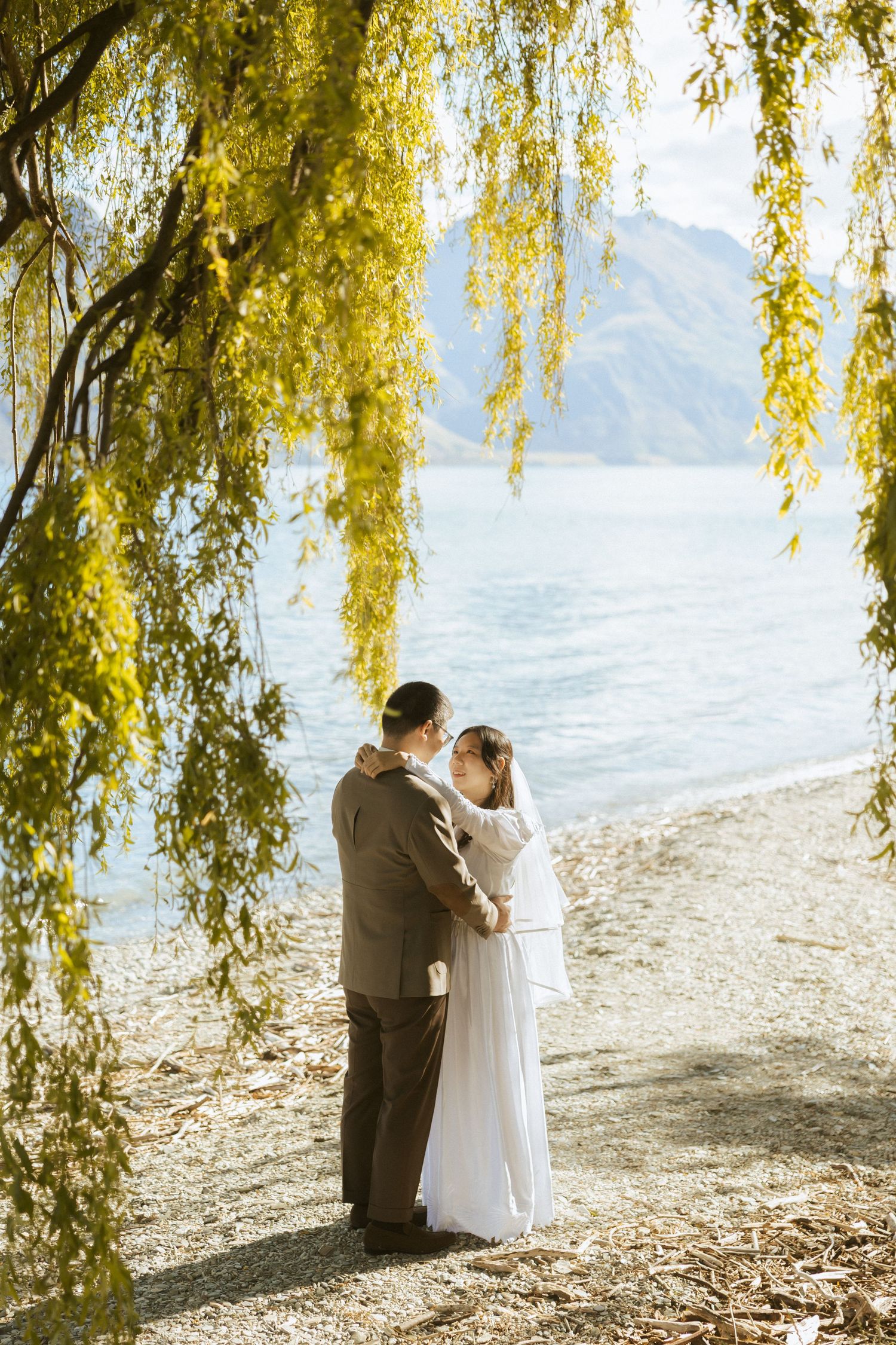 A couple shares an intimate moment under golden willow branches by a serene lakeside on their wedding day.