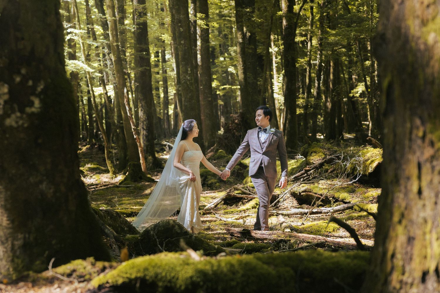 A couple in elegant white attire walks through a sunlit mossy forest during a romantic outdoor photo session.