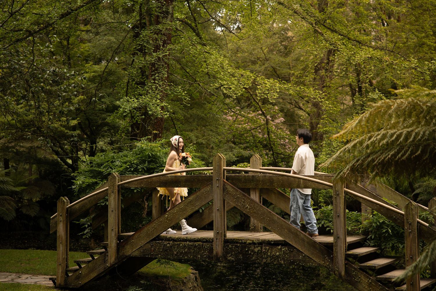 A scenic wooden footbridge spans across a peaceful garden setting surrounded by lush greenery and trees.