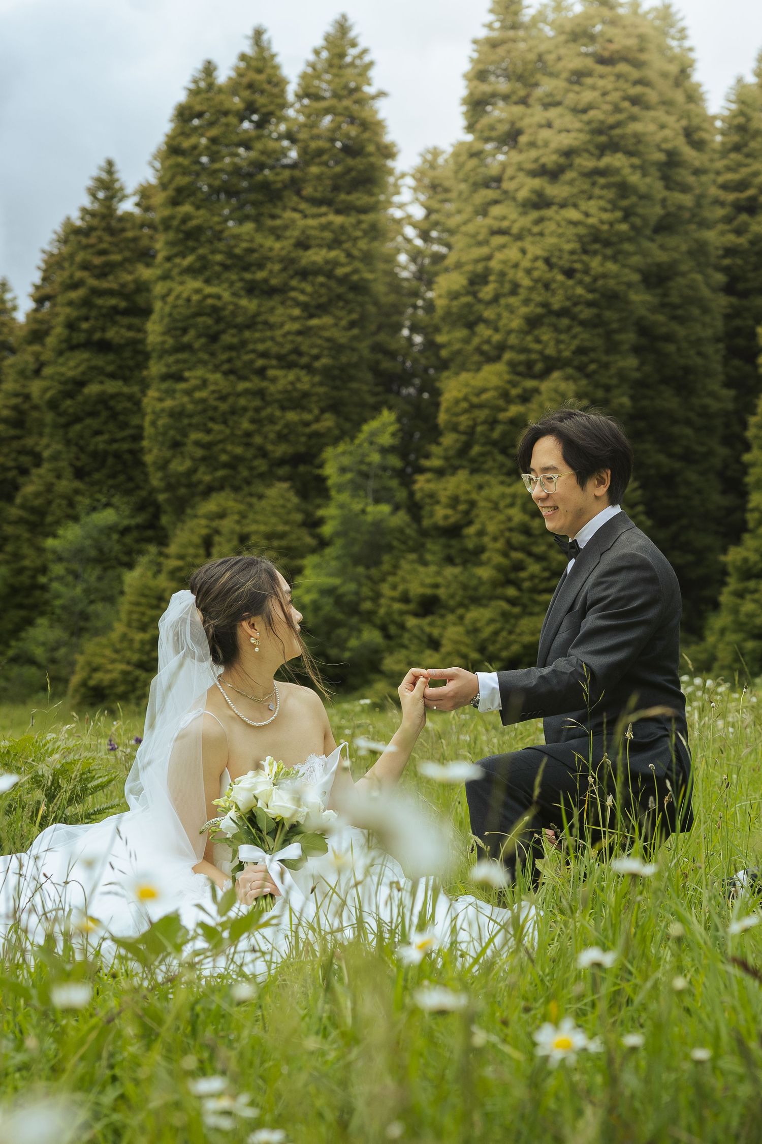 A romantic wedding photo in a meadow of daisies with evergreen trees in the background.