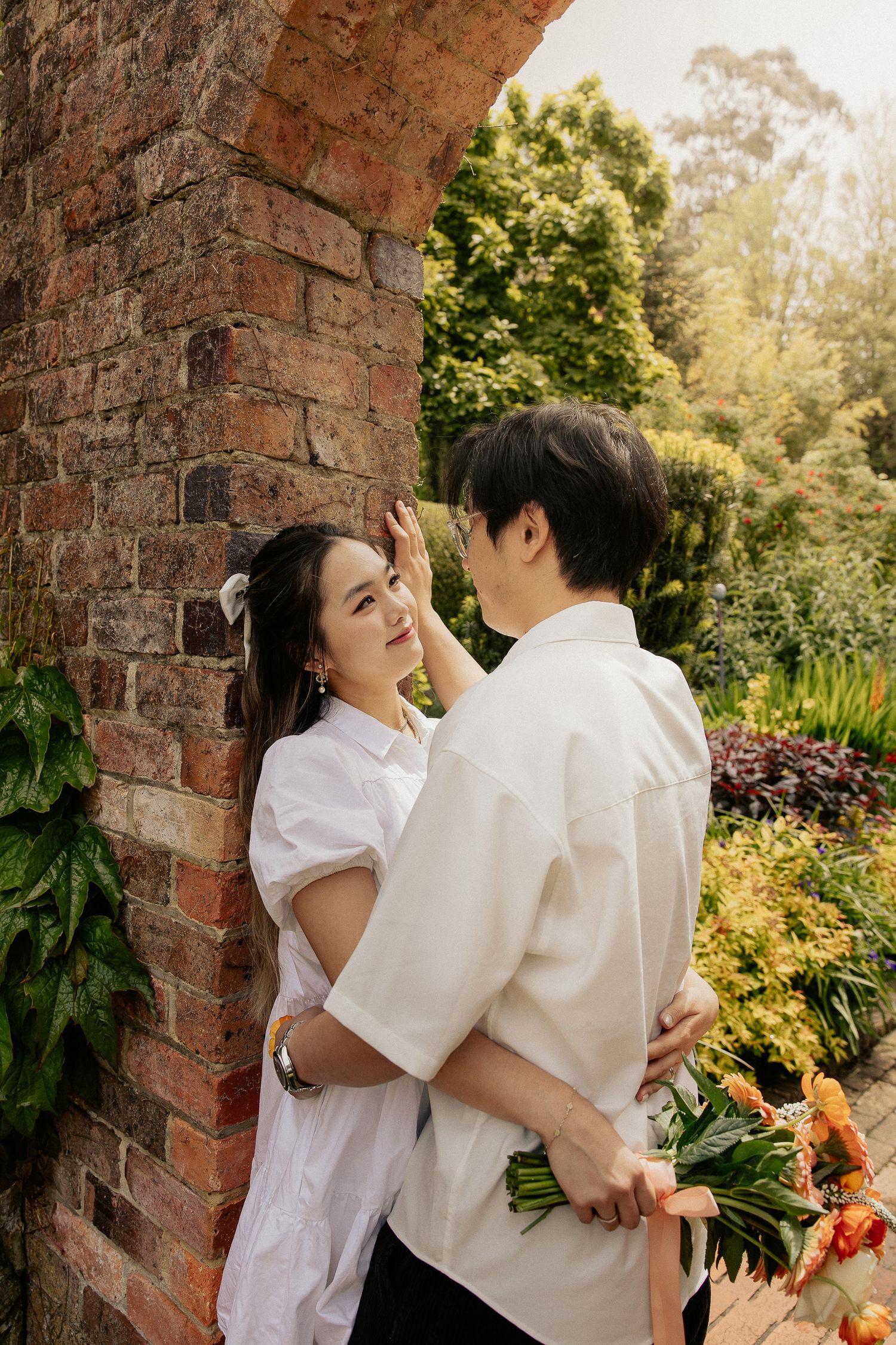 A couple in white outfits embrace romantically against a rustic brick wall in a lush garden setting.