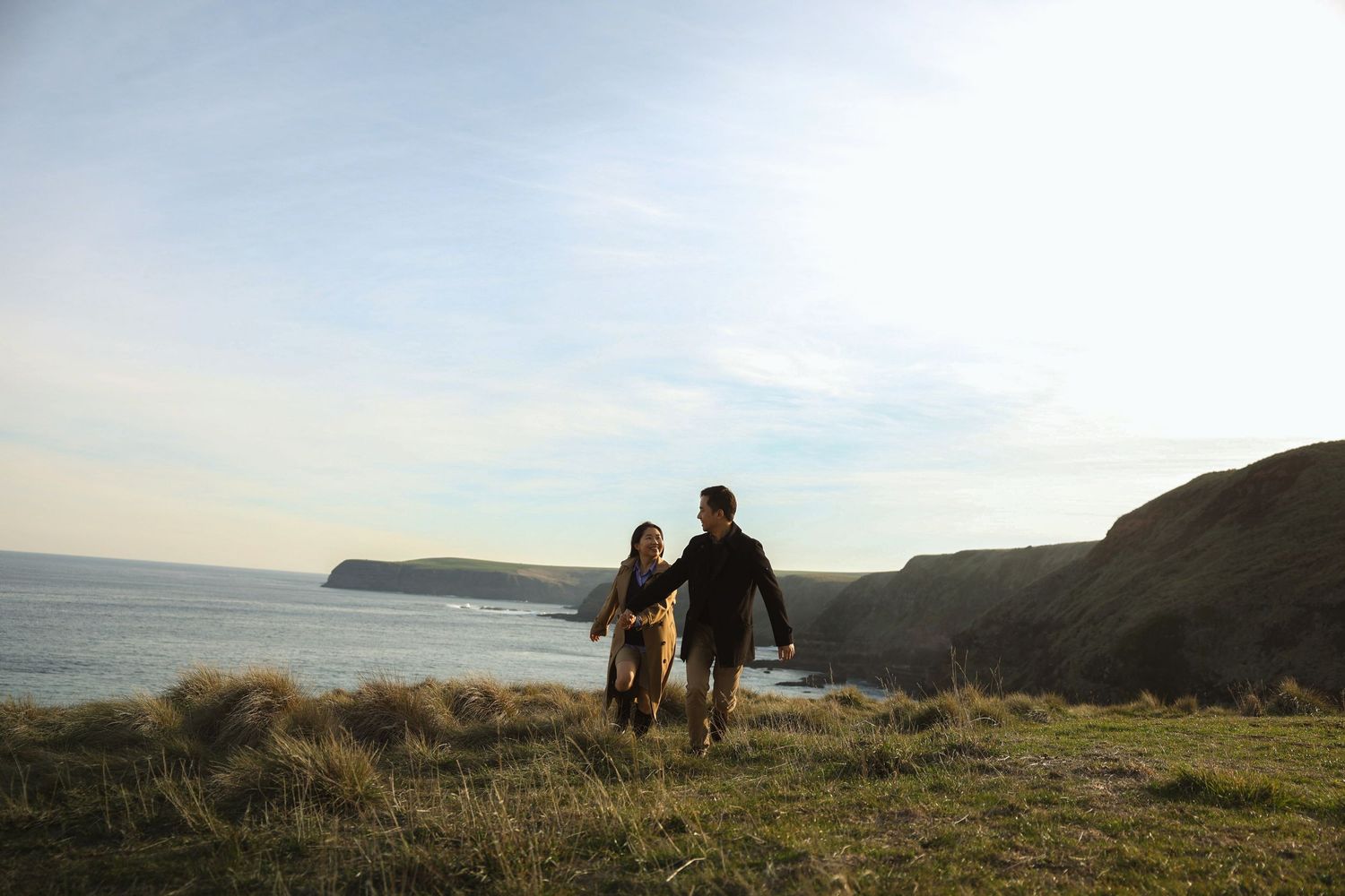 Two people walk together along a scenic coastal cliff path overlooking a calm ocean at sunset.