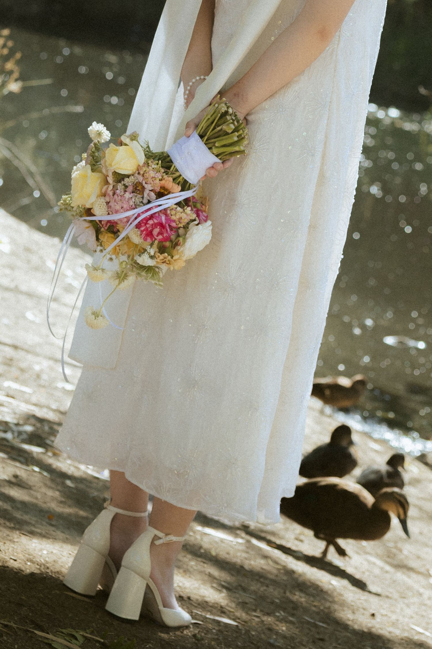 A bride in a white dress and heels stands near a pond with ducks visible in the background.