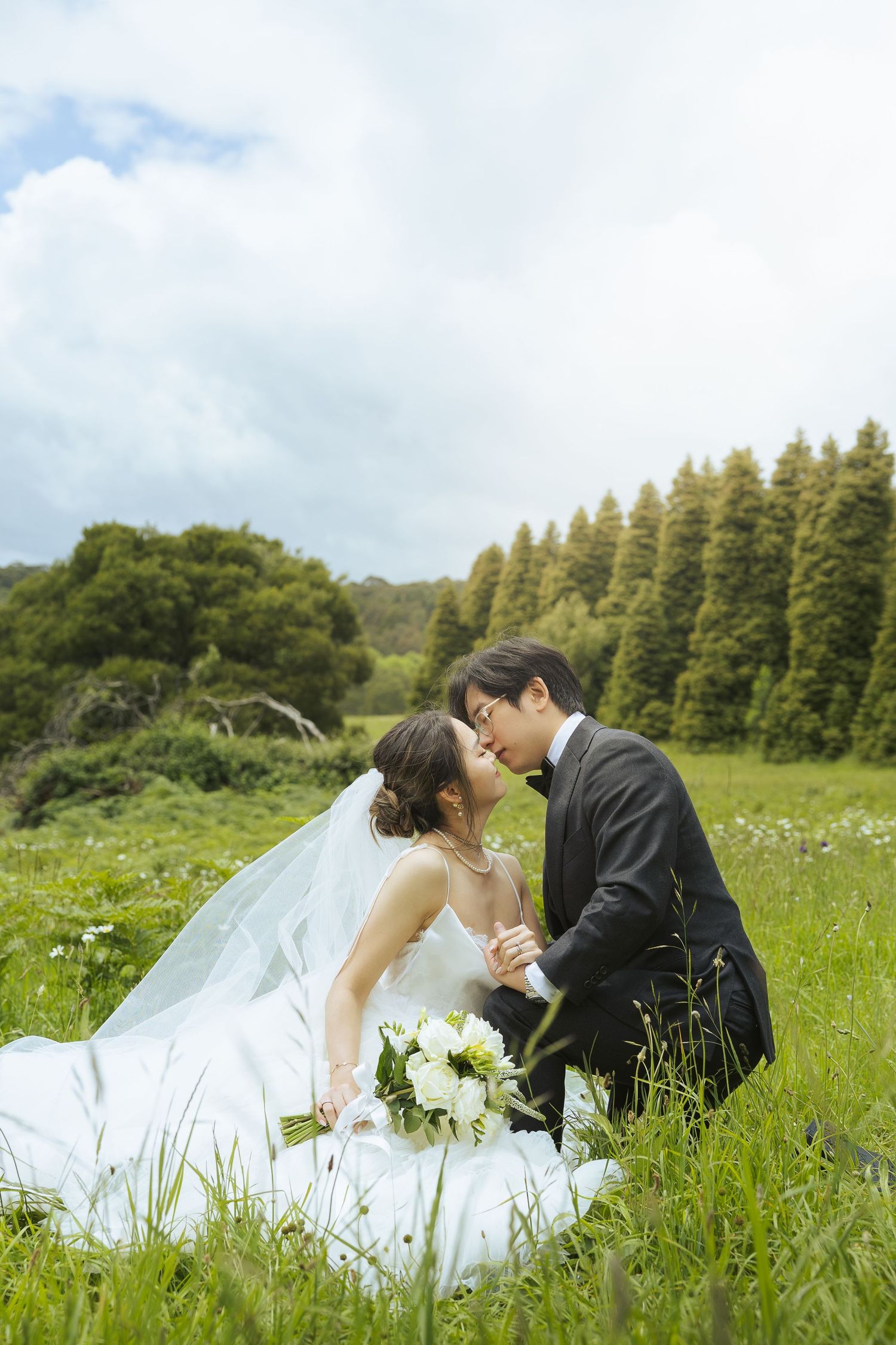 A romantic wedding photo sequence showing a couple laying in a green meadow with wildflowers and evergreen trees.