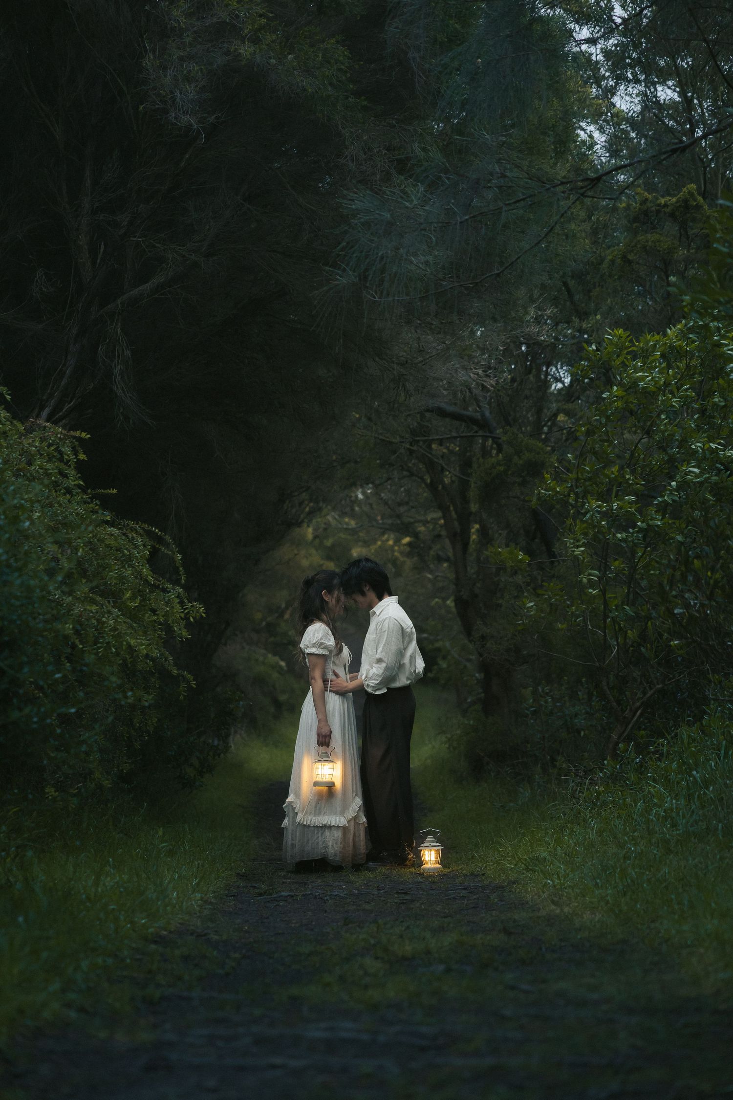 Two figures in vintage clothing walk along a dark forest path carrying a glowing lantern at dusk.