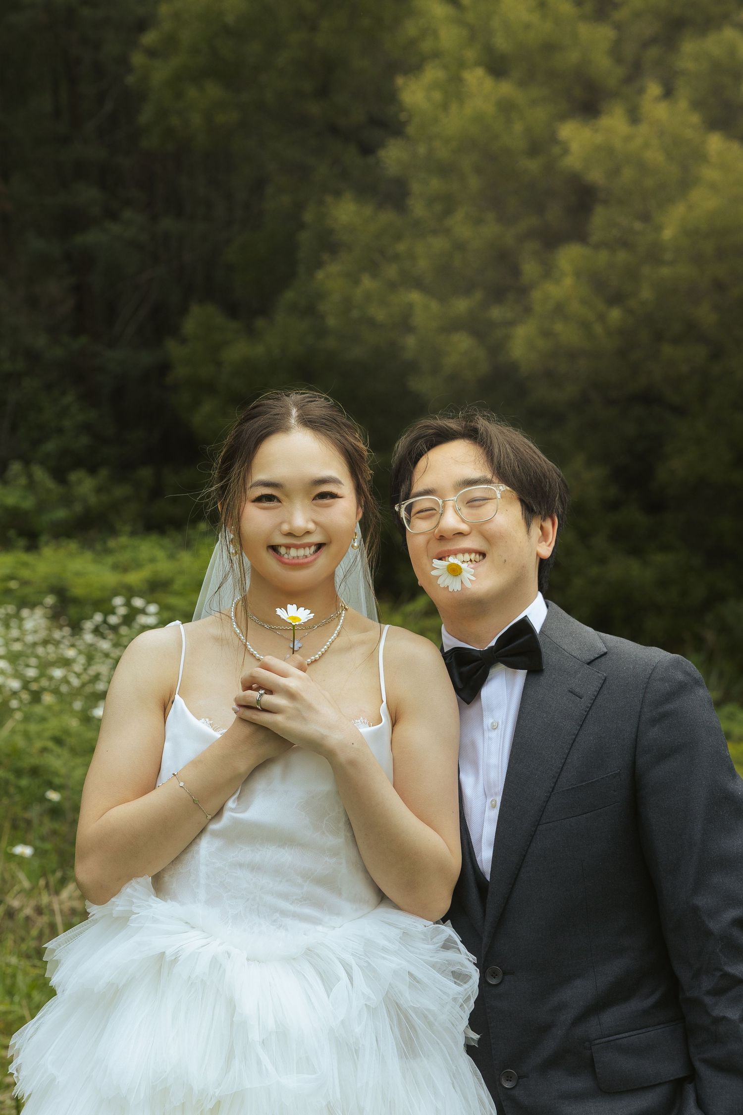 A couple in wedding attire poses outdoors with a small flower, surrounded by lush greenery.