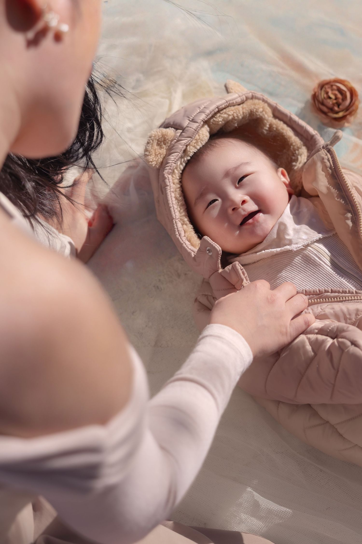 A small baby wearing a beige quilted onesie and hood lies on a light surface while being gently touched.