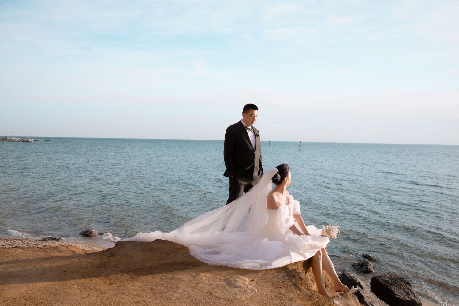 Couple in wedding attire embracing on a rocky beach at sunset with waves crashing in the background.