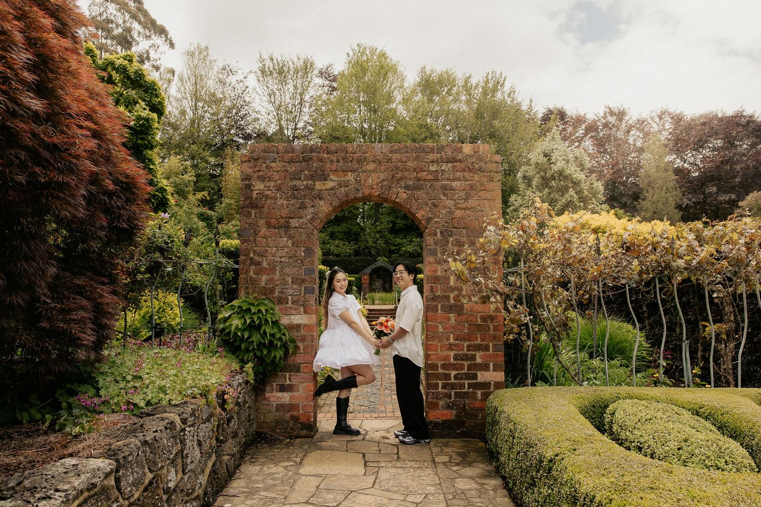 A wedding couple stands in a rustic stone archway surrounded by manicured gardens.