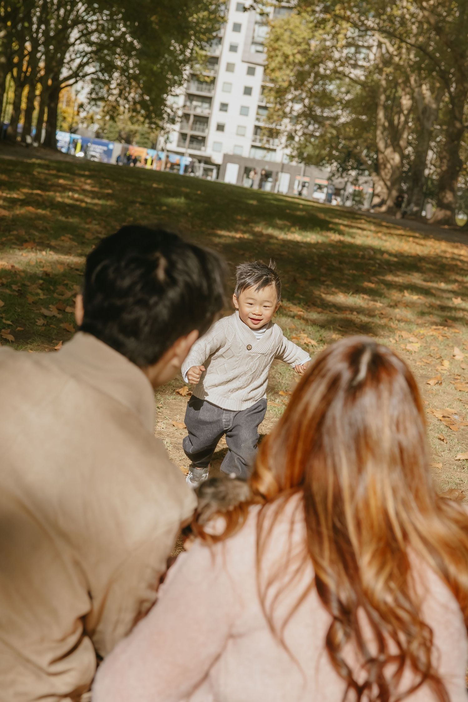 A toddler in a sweater runs joyfully through a park on an autumn day with apartment buildings in the background.
