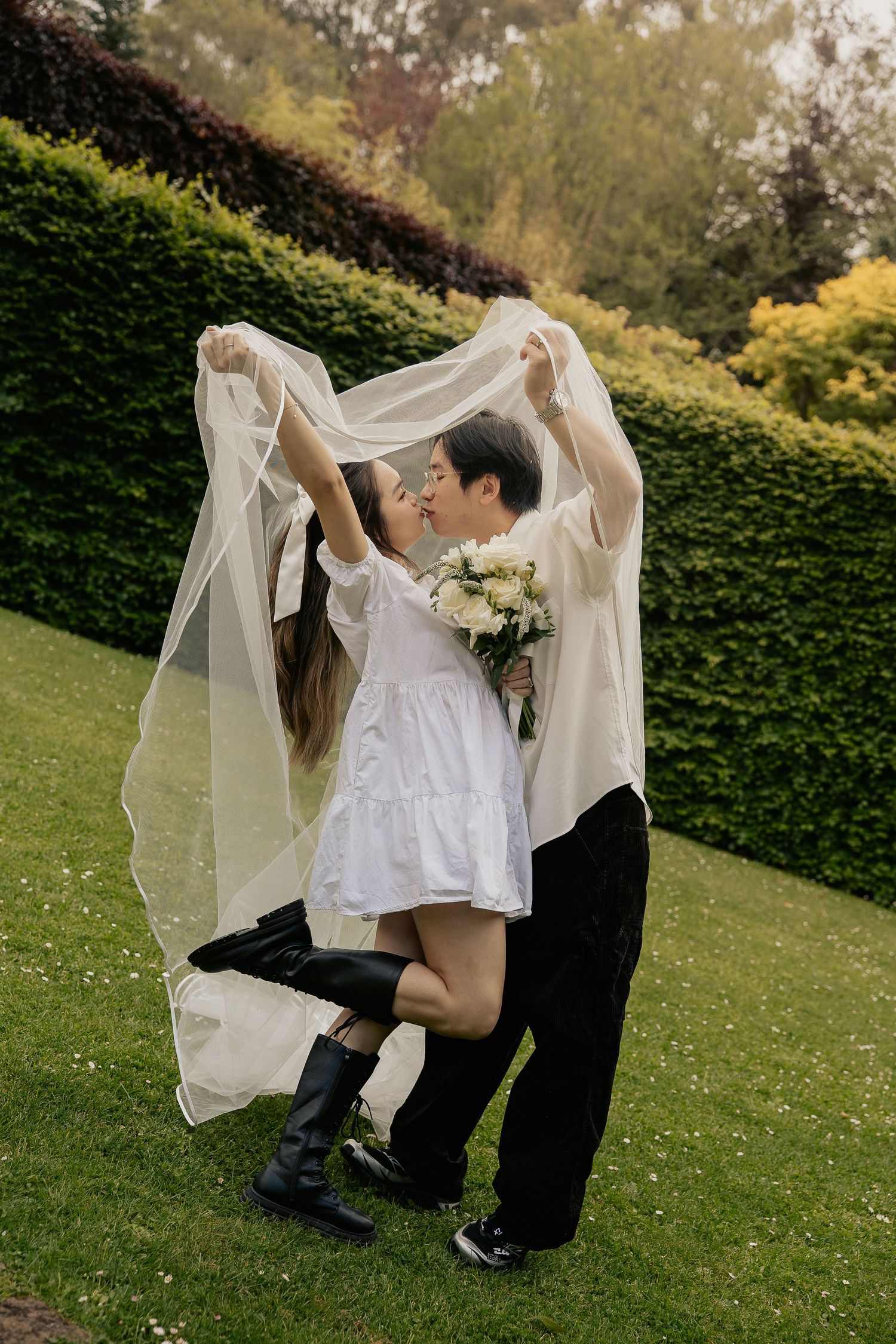 Children in white clothing play with a sheer fabric on a grassy lawn with a hedge backdrop.